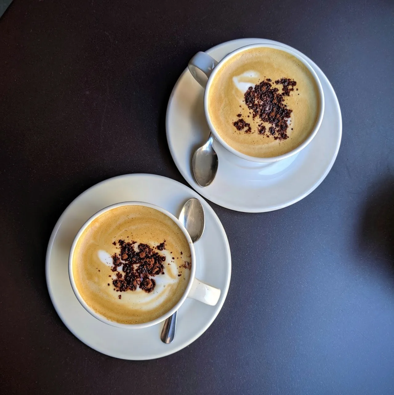 Two cups of cappuccino topped with cocoa on white saucers seen from above