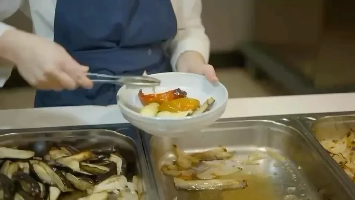 Chef plating roasted vegetables and sauce into a white bowl with tongs