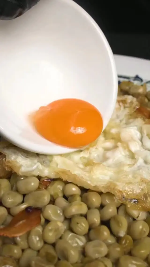 Pouring a raw egg yolk from a small bowl onto a fried egg over a plate of white beans