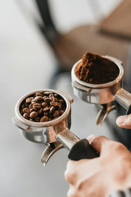 Two portafilters held side by side, one filled with whole coffee beans and one with ground coffee