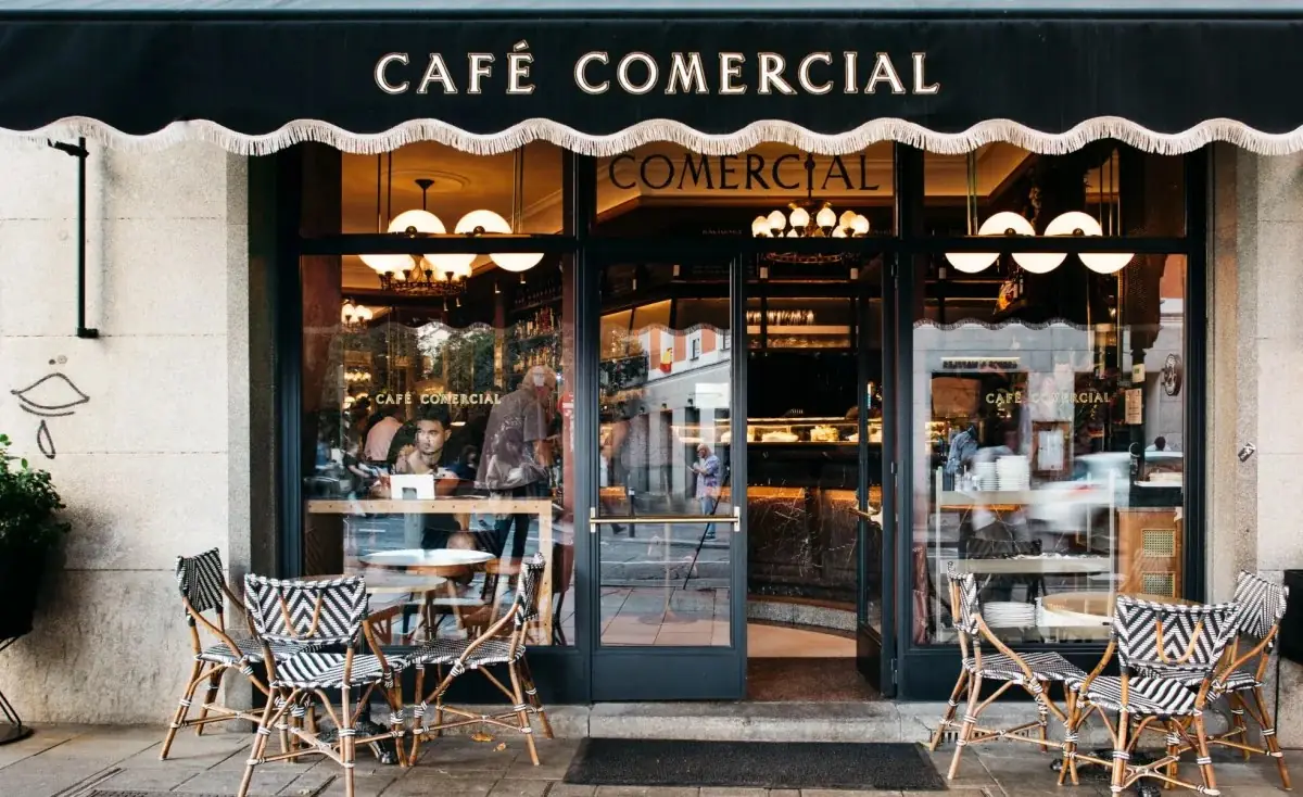 Cafe Comercial storefront with black awning reading CAFE COMERCIAL, glass frontage and rattan bistro chairs on the pavement