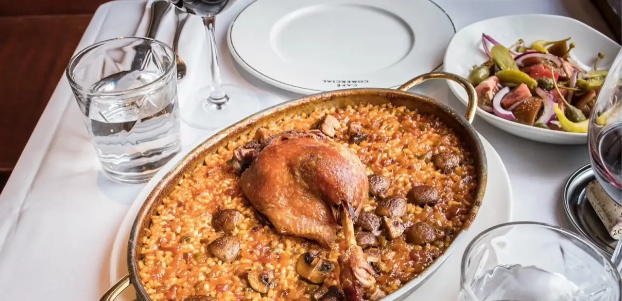 Rice dish with a confit duck leg and mushrooms in a metal pan, beside a pickle-and-onion salad and red wine on a white tablecloth