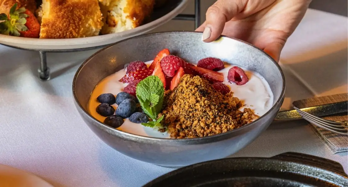 Yogurt bowl topped with strawberries, raspberries, blueberries, granola crumble and mint, served by a hand