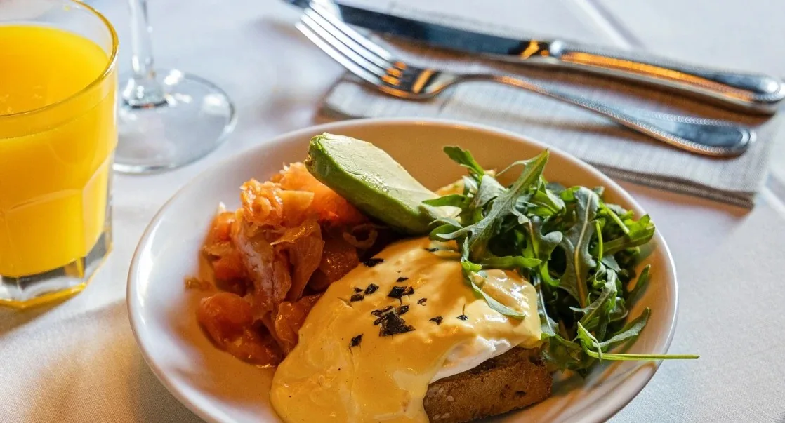 A plate with poached eggs and hollandaise, slices of avocado and rocket leaves, beside a glass of orange juice