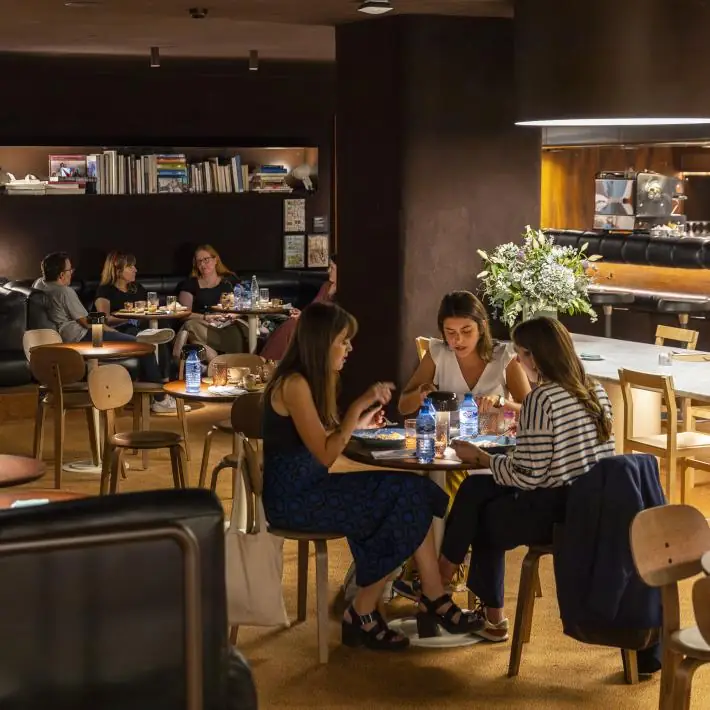 Dimly lit café dining room with diners at small round tables, leather banquettes, bookshelves and a marble counter