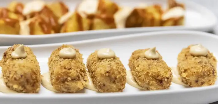 A row of five breaded croquetas each topped with a dot of aioli on a white plate, with patatas behind