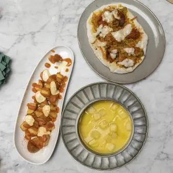 Three plated dishes on a marble table: patatas bravas, a topped tostada, and a bowl of soup
