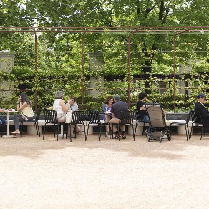 Café terrace with diners at tables against a green trellis of climbing plants, gravel floor in foreground