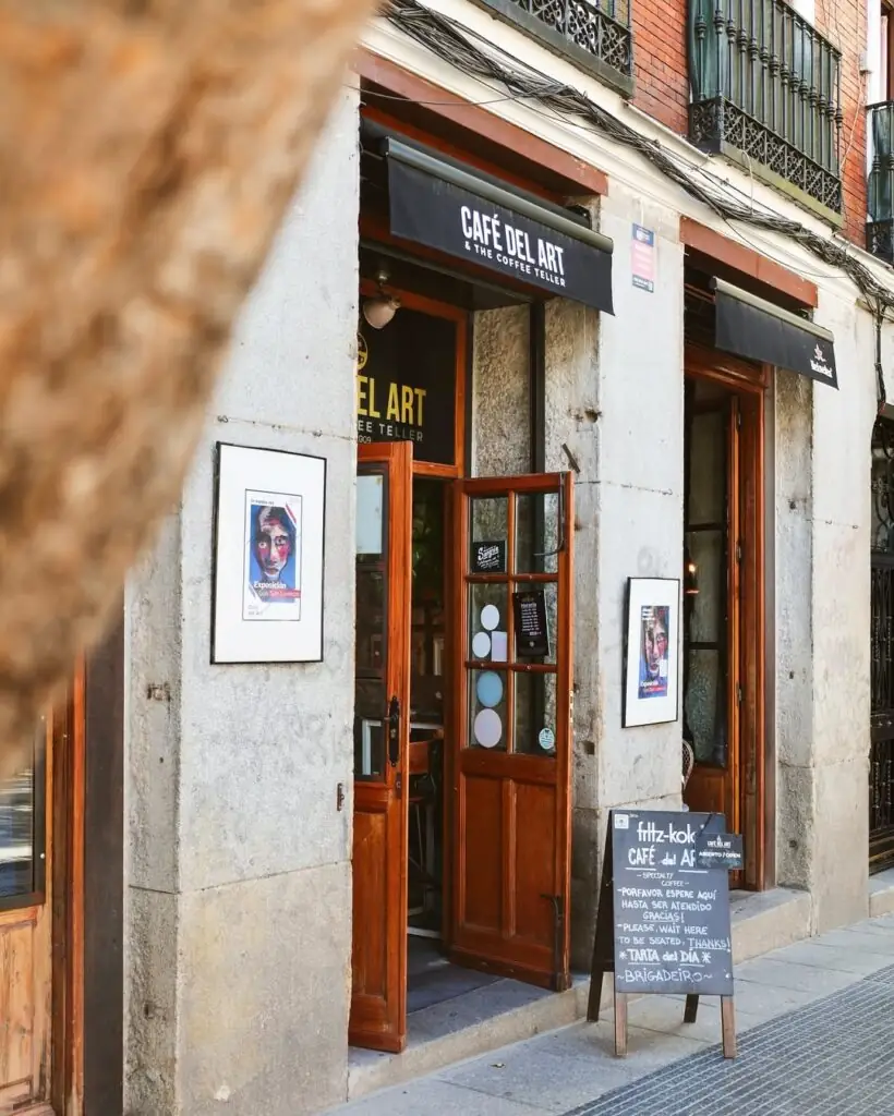 Street storefront of Cafe del Art & The Coffee Teller, with black awning signage, a wooden door, a chalkboard sign and framed artwork on a stone facade