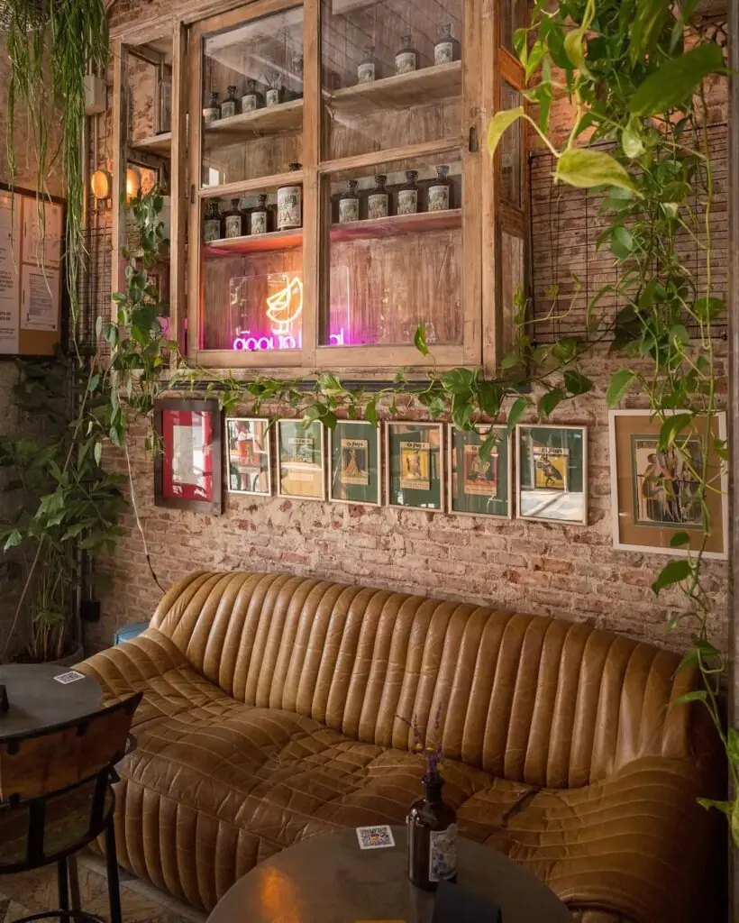 Cosy cafe interior with a tan leather sofa, exposed brick wall, hanging plants, a glazed apothecary cabinet with a neon sign and framed prints