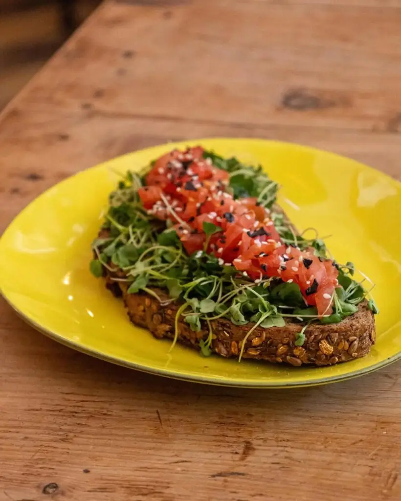Slice of multigrain seeded toast topped with microgreens, diced tomato and sesame on a yellow plate