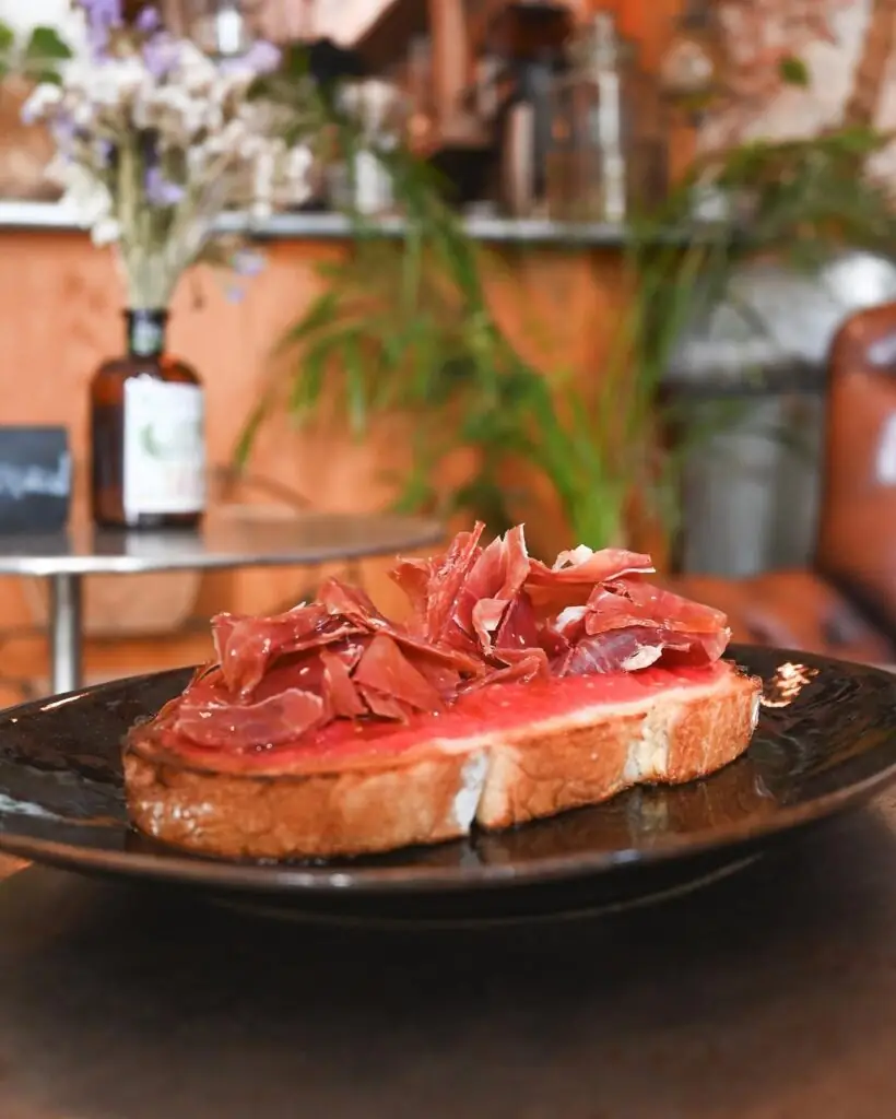 Tostada of rubbed tomato and Iberian ham on toasted bread on a dark plate, with a warm café interior behind
