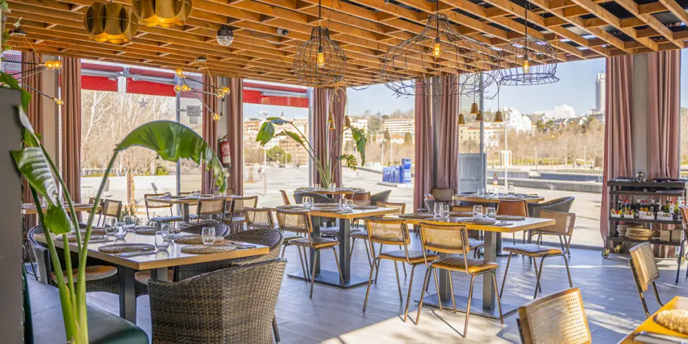 Terrace dining room with set tables, rattan chairs, wooden slat ceiling with cage pendant lamps and open windows
