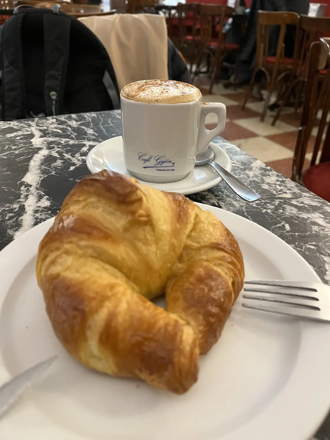 A flaky croissant on a white plate beside a cappuccino dusted with cocoa, served in a cup marked 'Cafe Gijon' on a dark marble table