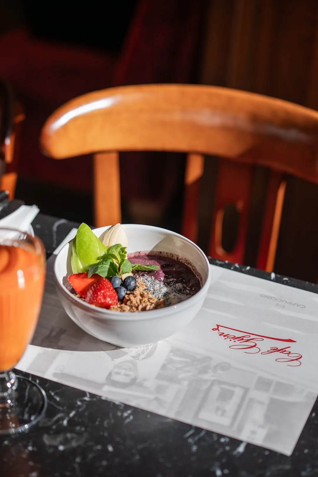 A smoothie bowl topped with strawberry, blueberries, apple slices, granola and mint, next to a glass of orange juice on a marble table