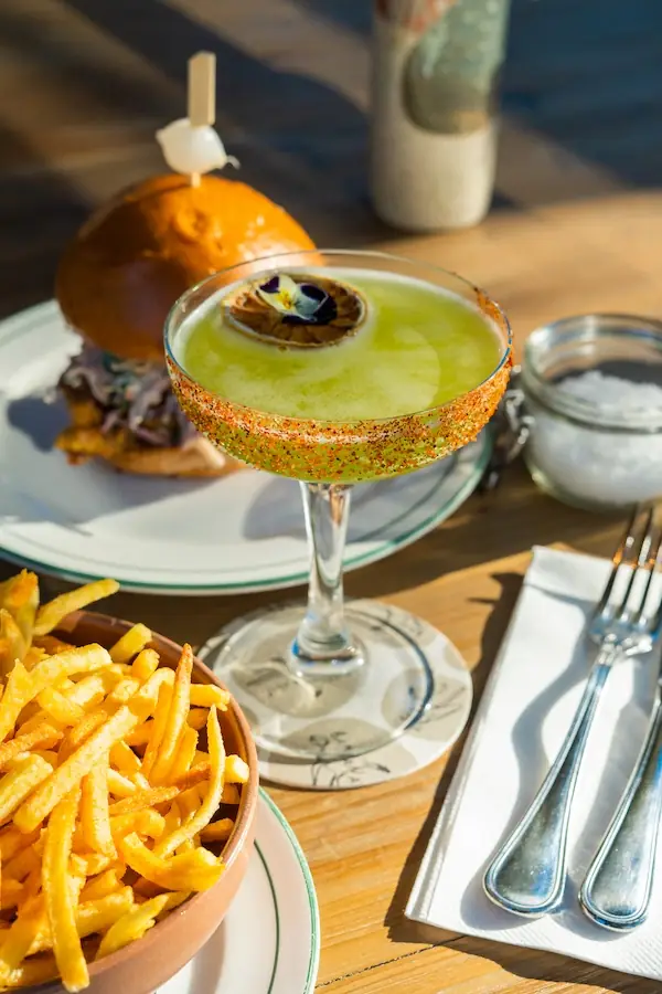 A burger with fries and a green cocktail on a sunlit table at Café Poupette