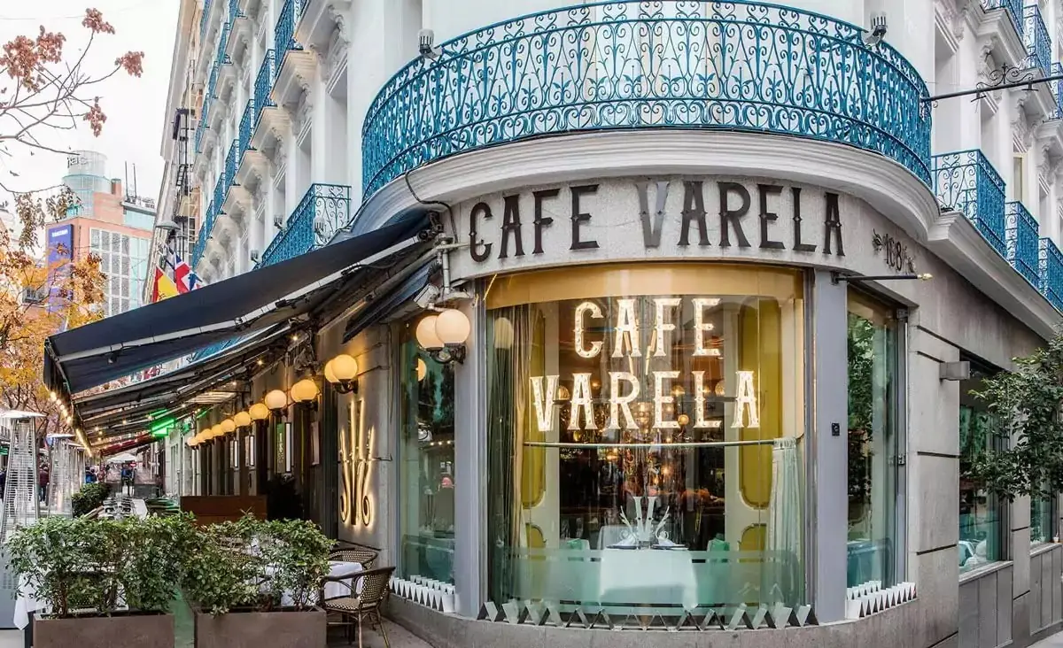 Corner exterior of Cafe Varela with an illuminated CAFE VARELA sign in the curved window, a blue wrought-iron balcony above and a terrace awning