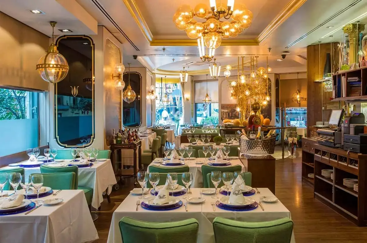 Ornate dining room with white-clothed tables set with blue chargers, green upholstered chairs, gold globe chandeliers and mirrored walls