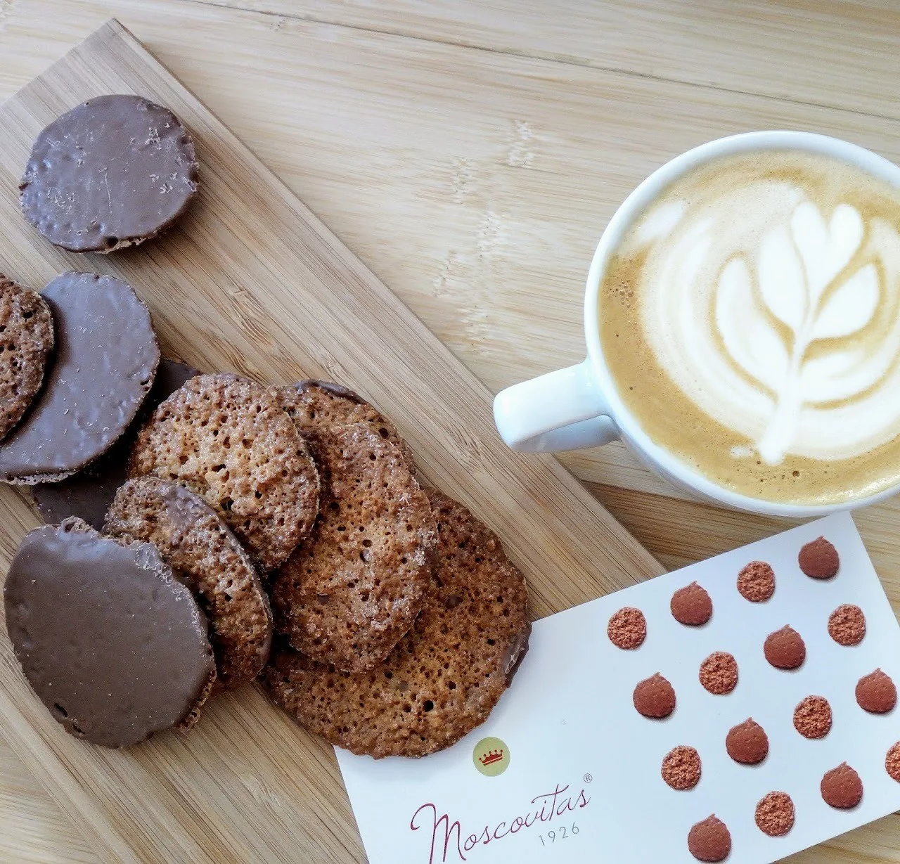 A latte with art in a white cup beside dark chocolate biscuits on a wooden board