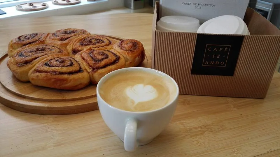 Two cinnamon rolls on a wooden board next to a cappuccino with heart-shaped foam art
