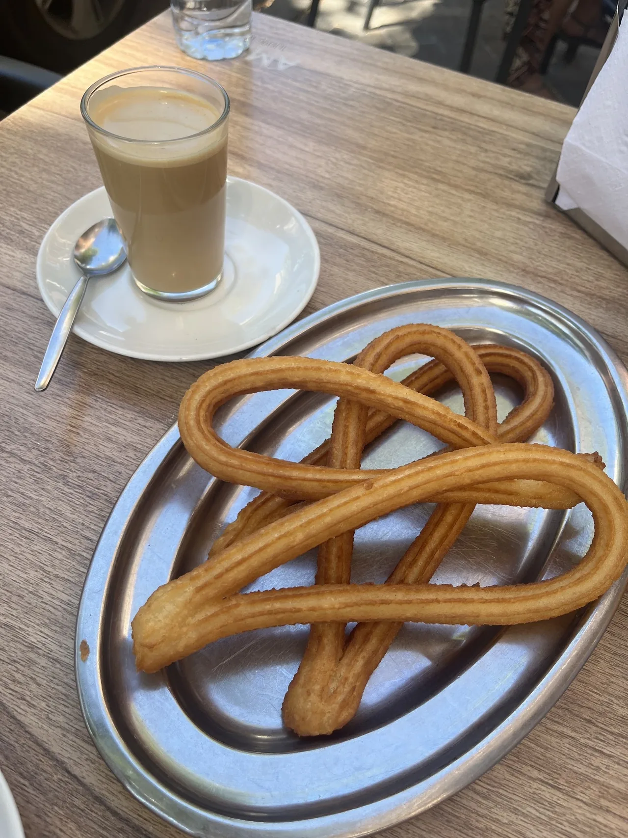 A looped churro on a metal tray beside a glass of coffee with milk