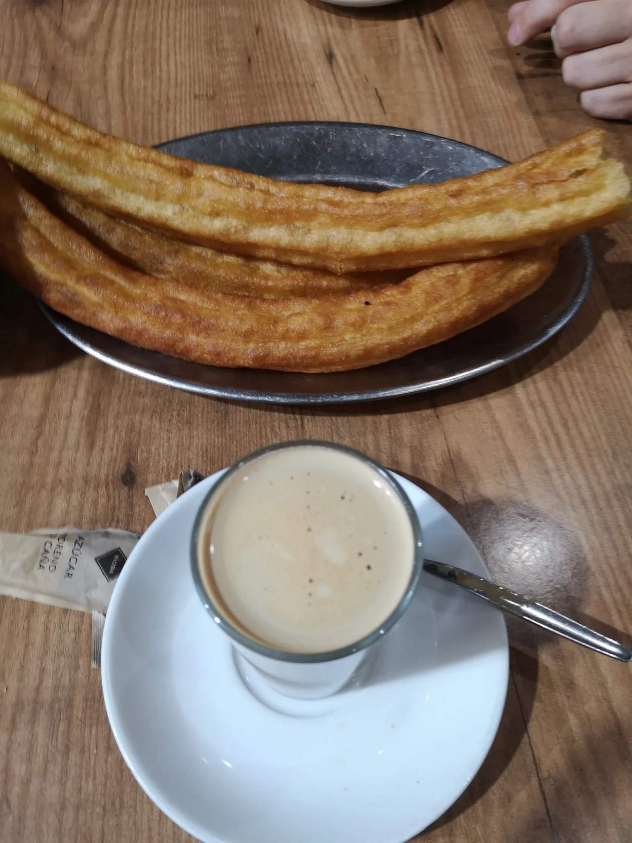 Long churros on a metal tray above a glass of coffee on a saucer