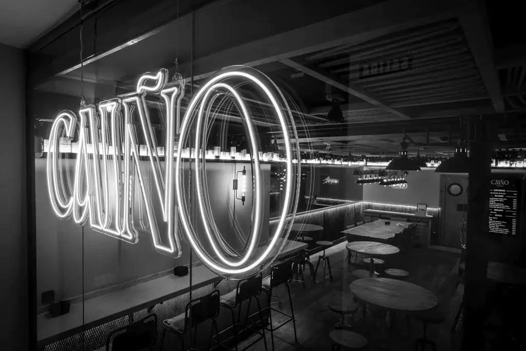 Black-and-white view through a glass front of the venue interior with a large neon 'Caiño' sign, backlit wine wall, wooden tables and stools
