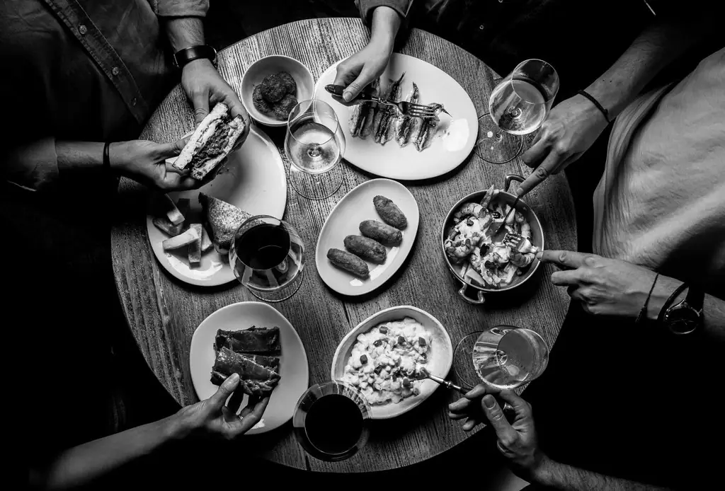 Black-and-white overhead of a shared table with fried croquetas, marinated anchovies, an ensaladilla, cured meat and glasses of wine, diners' hands reaching in