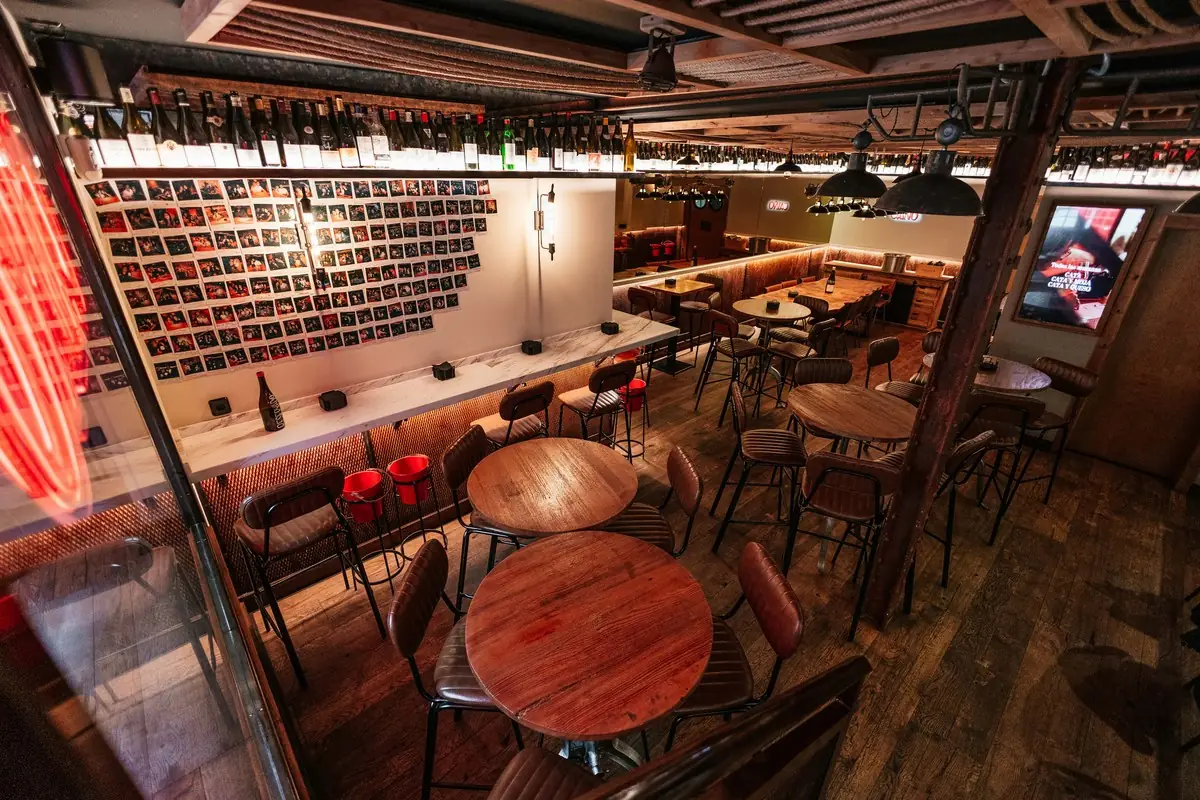 Interior of the wine bar with wooden tables, red stools, and a wall of wine bottles.