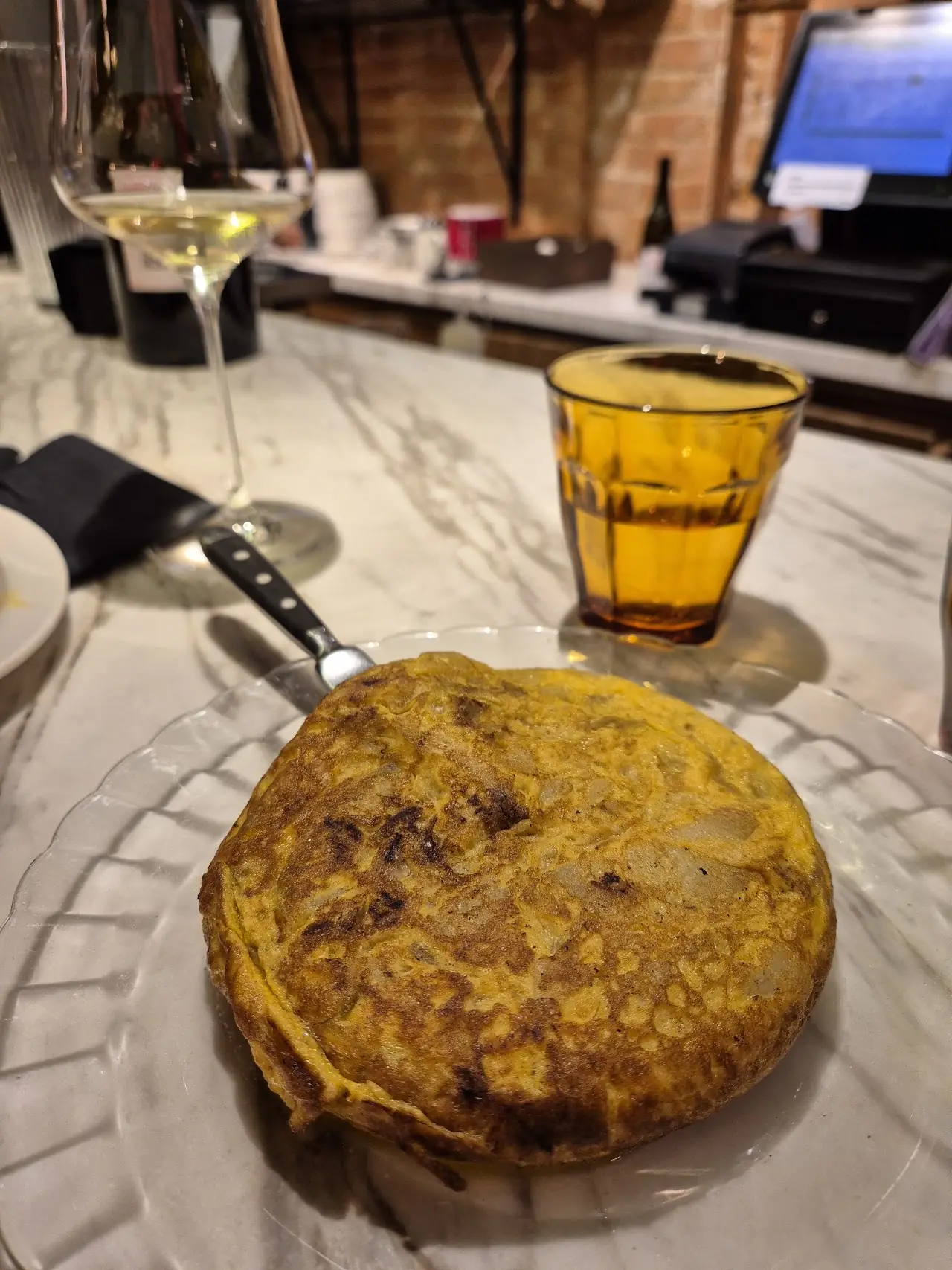 A round Spanish tortilla on a plate beside a glass of white wine and an amber drink.