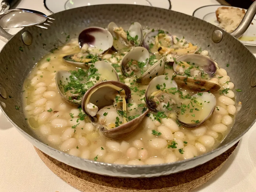 A metal pan of open clams over white beans, garnished with chopped parsley