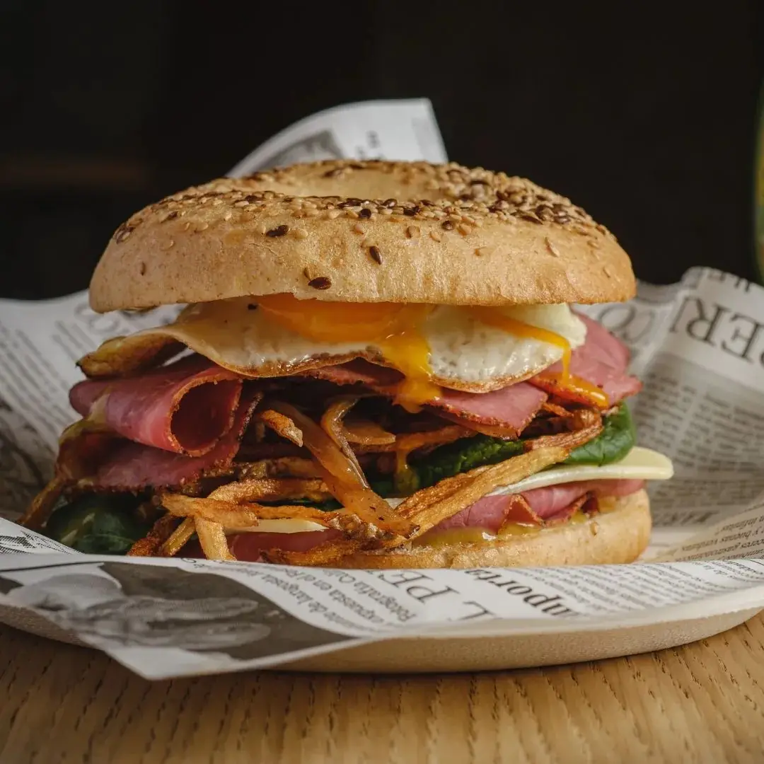 Sesame bagel sandwich stacked with fried egg, cured ham, spinach, cheese, and crispy fried onion strings, served on newspaper-lined paper at Cal Marius 449