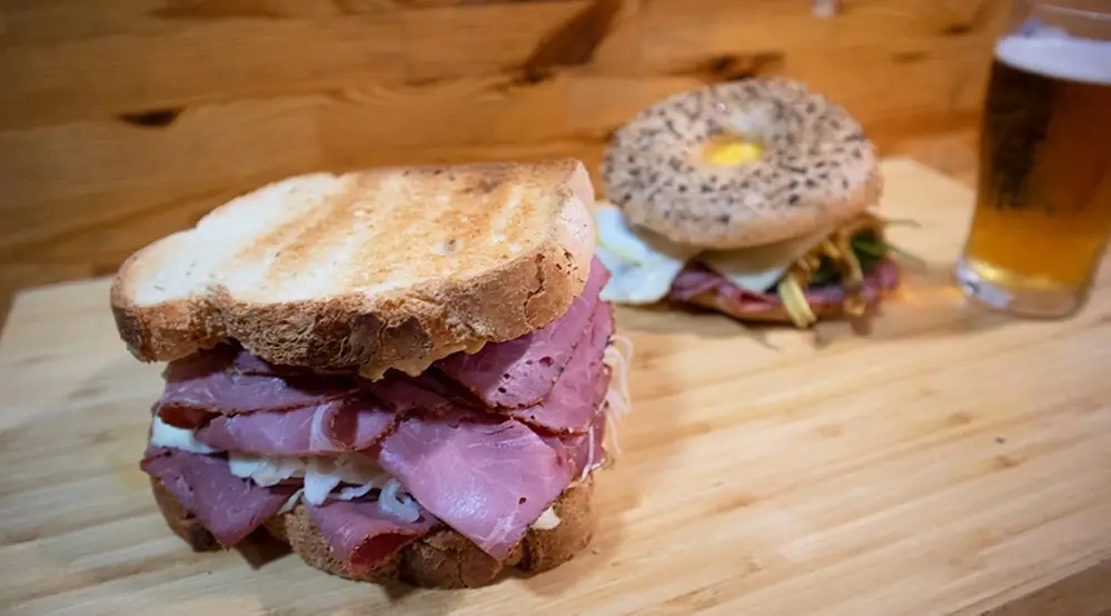 Toasted sandwich stacked with sliced cured meat on a wooden board, with a sesame bagel sandwich and a glass of beer in the background at Cal Marius 449