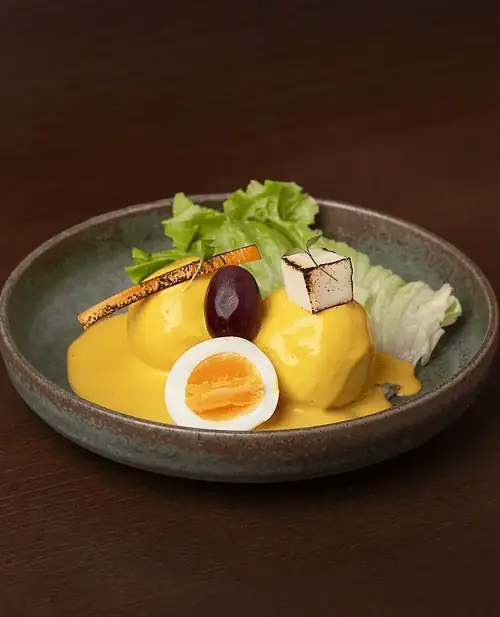 Papa a la huancaina: boiled potato under yellow cheese sauce with a halved soft egg, black olive, lettuce and a grilled skewer on a ceramic bowl over a dark table