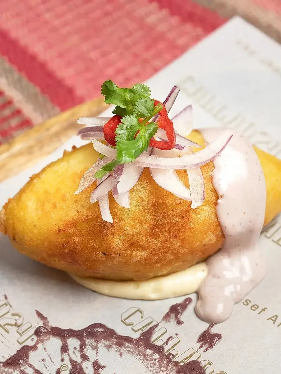 A golden fried potato dumpling topped with red onion and sauce on a printed paper mat