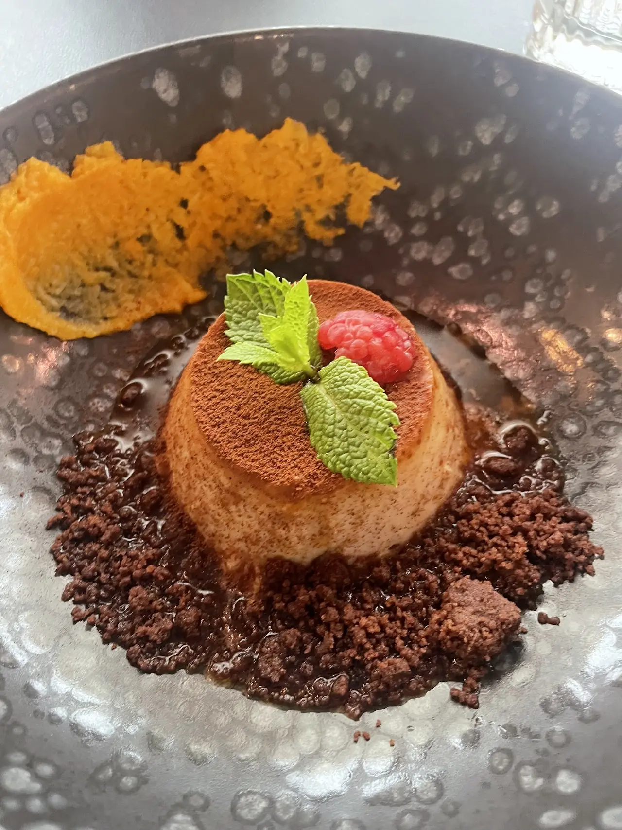 A round dessert dusted with cocoa powder, topped with mint and a raspberry, on a bed of chocolate crumble.