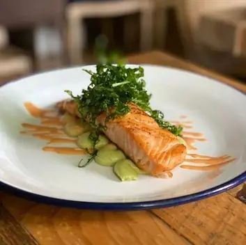 Seared salmon fillet topped with shredded greens over pureed dots and a drizzle of orange sauce on a white enamel plate