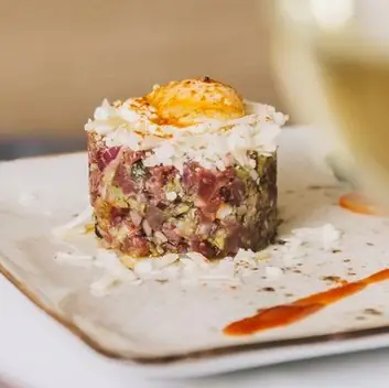 Tuna tartare timbale topped with an egg yolk and grated cheese on a beige plate with a smear of orange sauce