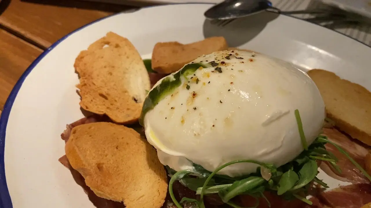 A burrata cheese ball topped with black pepper, served with toasted bread slices and cured ham