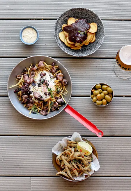 Overhead spread of tapas plates on a wooden table, including fried fish, octopus with potatoes, a dish in dark sauce, olives, and beer, at Camping Mar