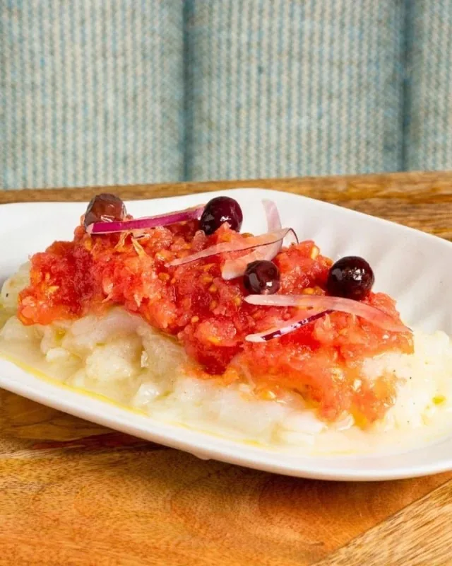 A white fish fillet on a white dish under grated fresh tomato, slivered red onion and black olives, dressed with olive oil, on a wooden table