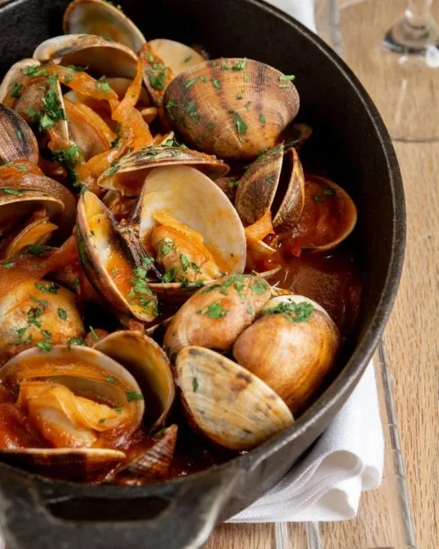 Clams opened in a tomato and parsley sauce in a black cast-iron pan, set on a white napkin on a wooden table