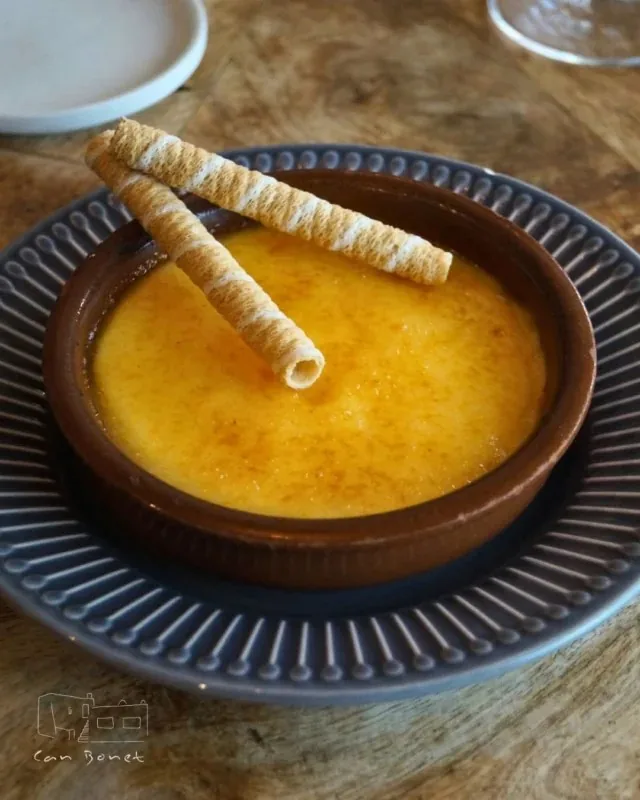 A caramelised custard in a brown earthenware cazuela on a ribbed grey plate, two rolled wafer biscuits laid across it