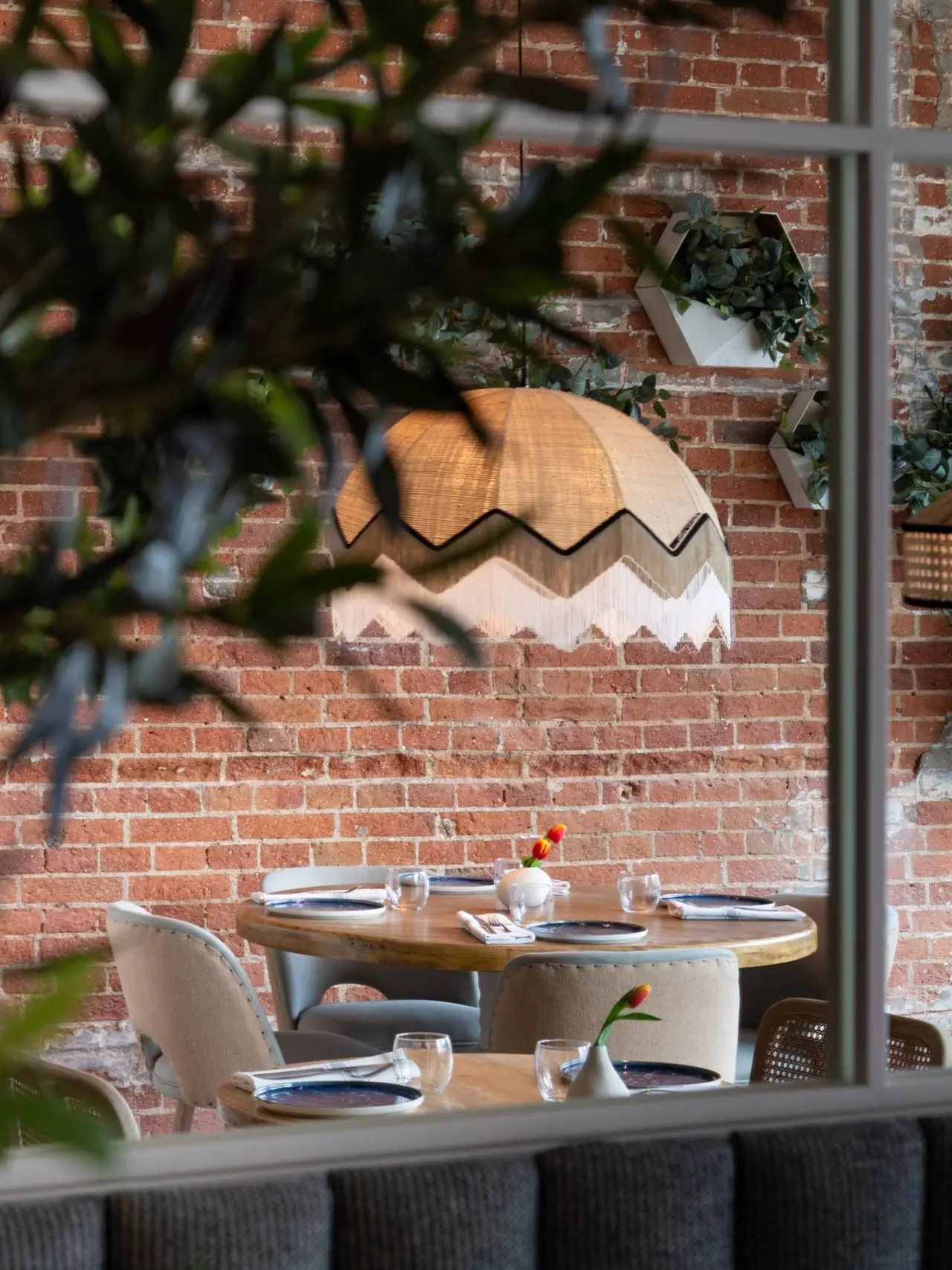 Dining room interior with an exposed brick wall, a woven pendant lamp, and a table set with blue-rimmed plates and glassware