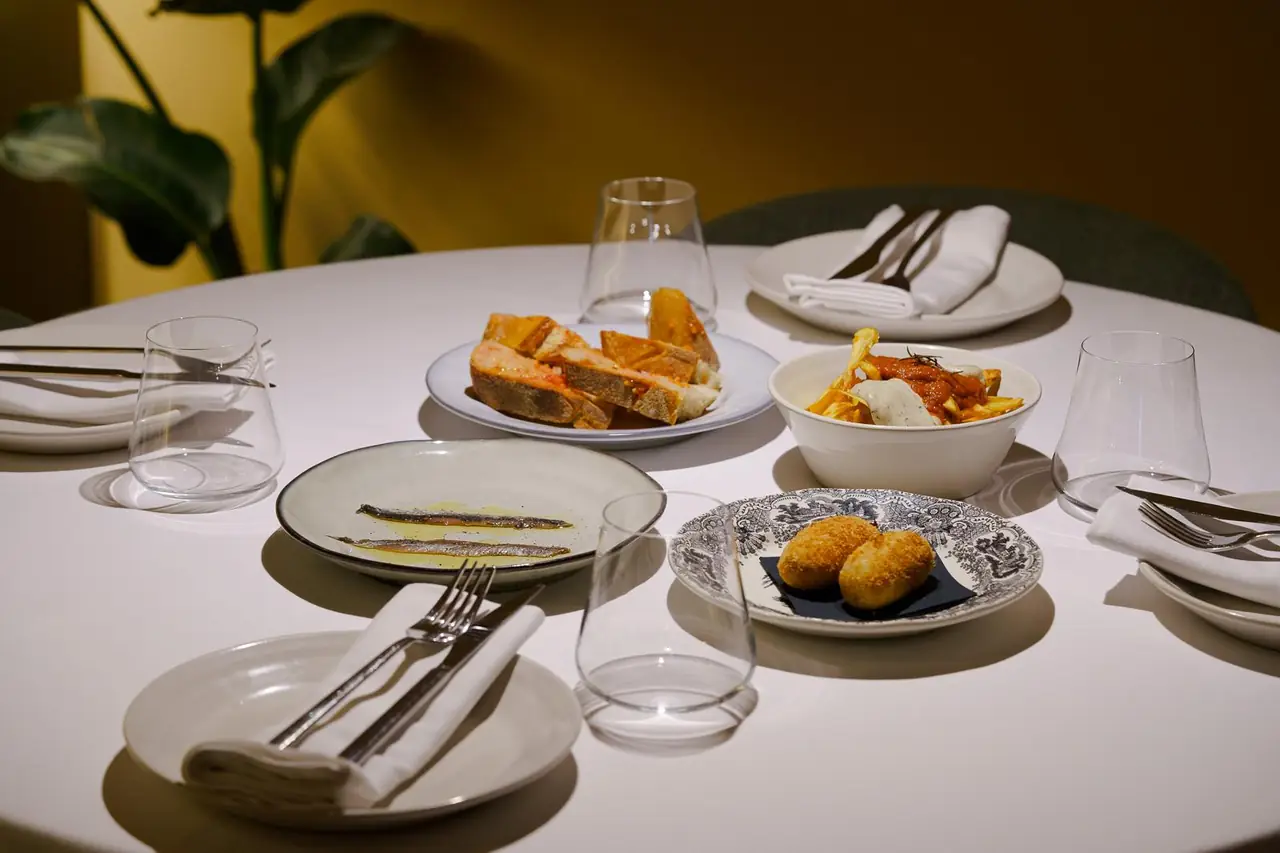 Table spread of tapas at Can Ugal: pa amb tomàquet with anchovies, a bowl of patatas bravas with sauce, a plate of two whole anchovies in oil, and two croquettes on a decorative plate