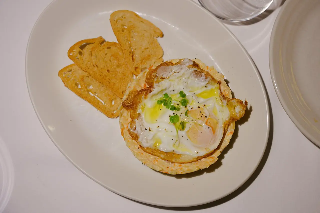 Overhead shot of a fried egg on a savory potato-carrot base topped with olive oil and microgreens, served with toasted bread slices at Can Ugal