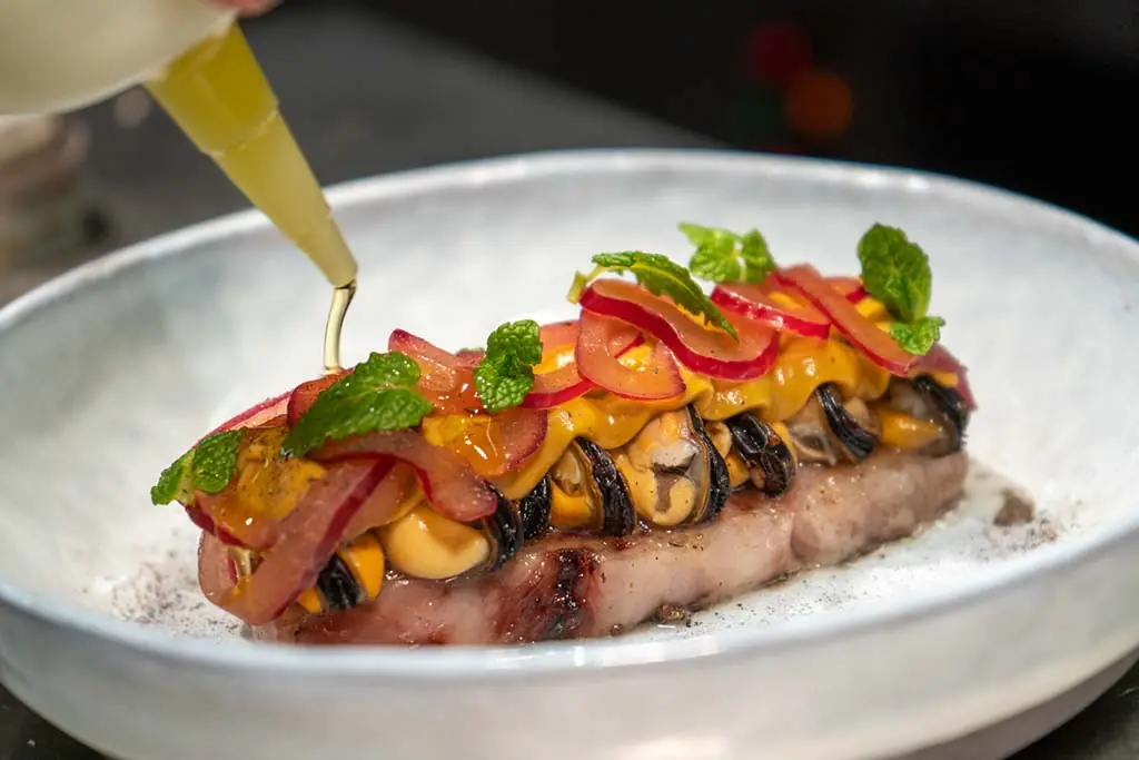 Grilled fish topped with charred mussels, pickled red onion, mango puree, and mint leaves, finished with a citrus dressing being poured over the top, at Capet.