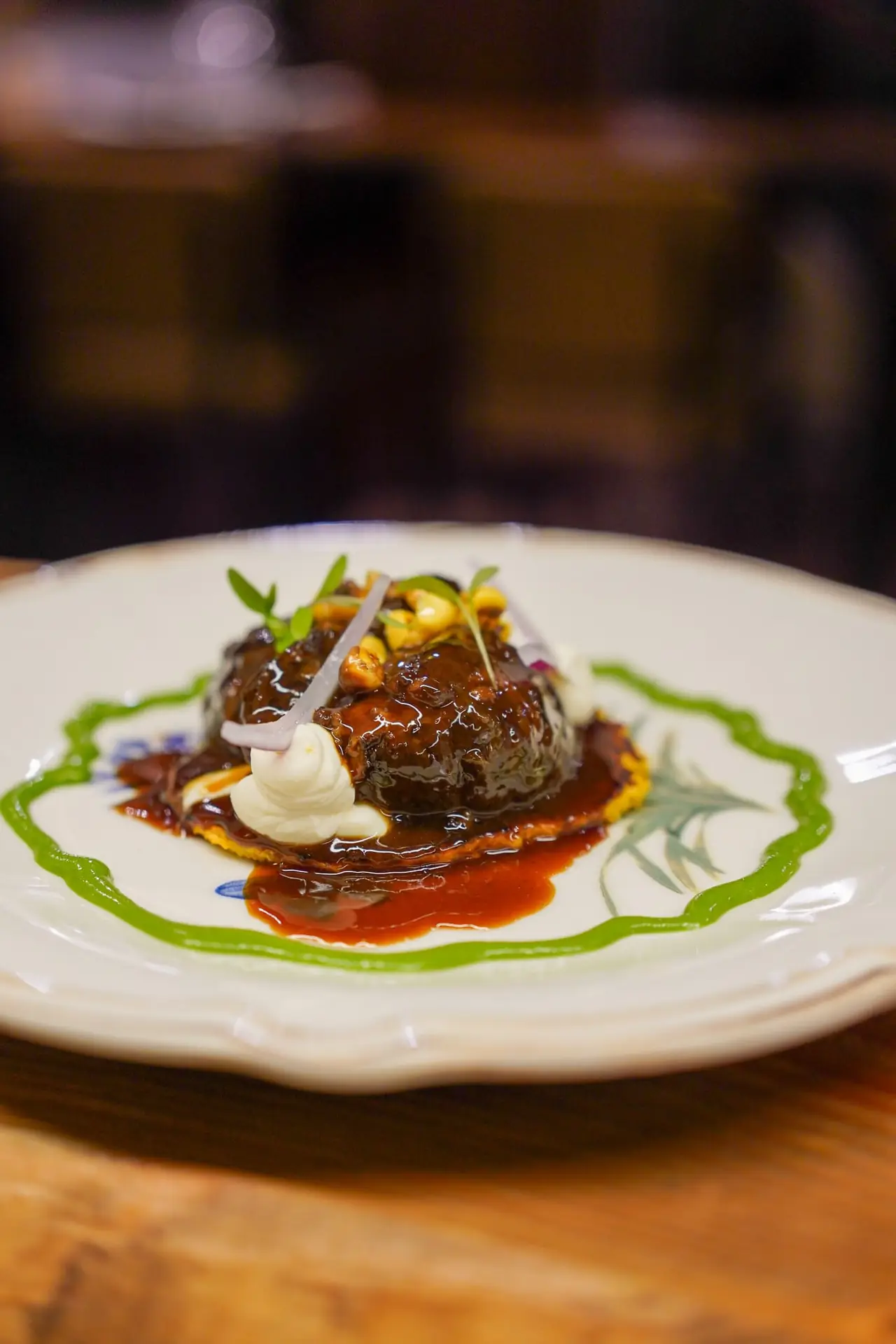 Braised meat in a dark glossy sauce over a corn base, with a red sauce swirl, whipped cream quenelle, pickled red onion, and a green herb sauce ring on a decorative plate, at Capet.