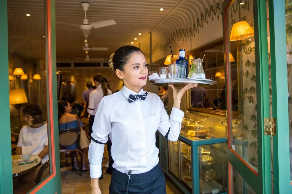 A waitress carrying a tray through the café doorway past the cake counter