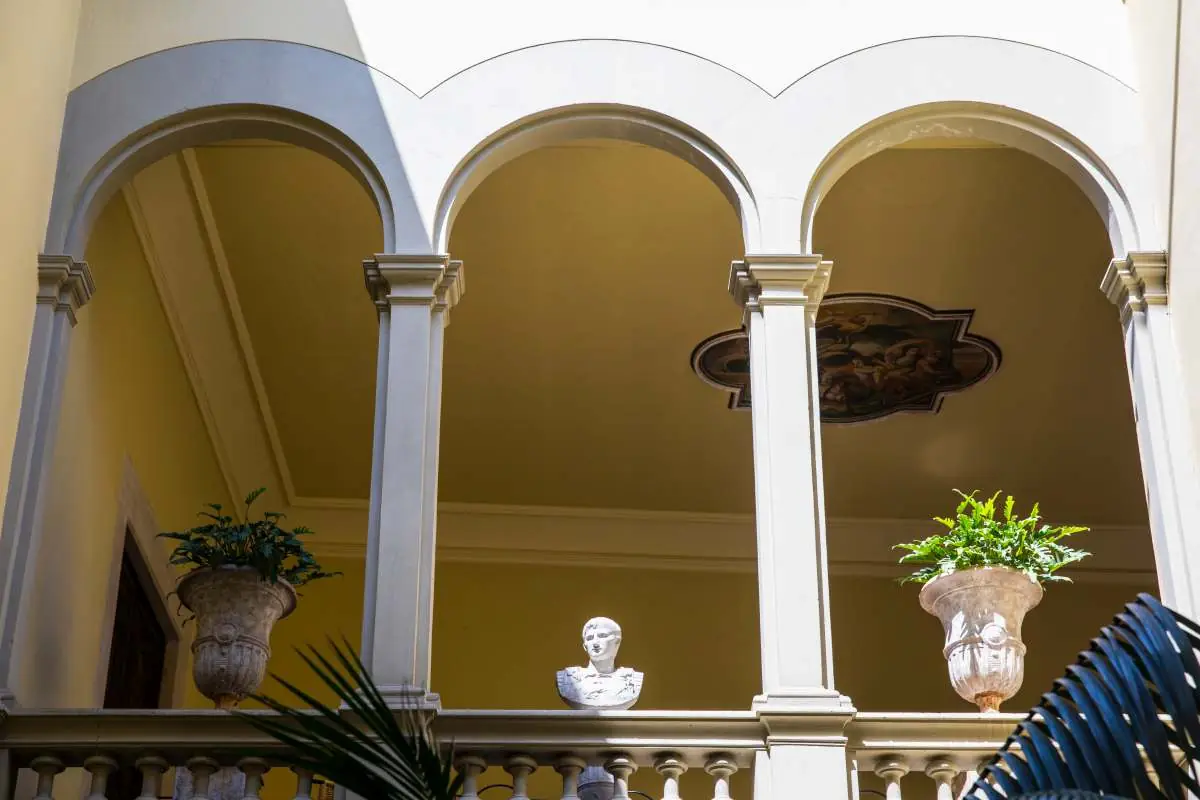 The arched upper gallery of the courtyard with plants and a marble bust